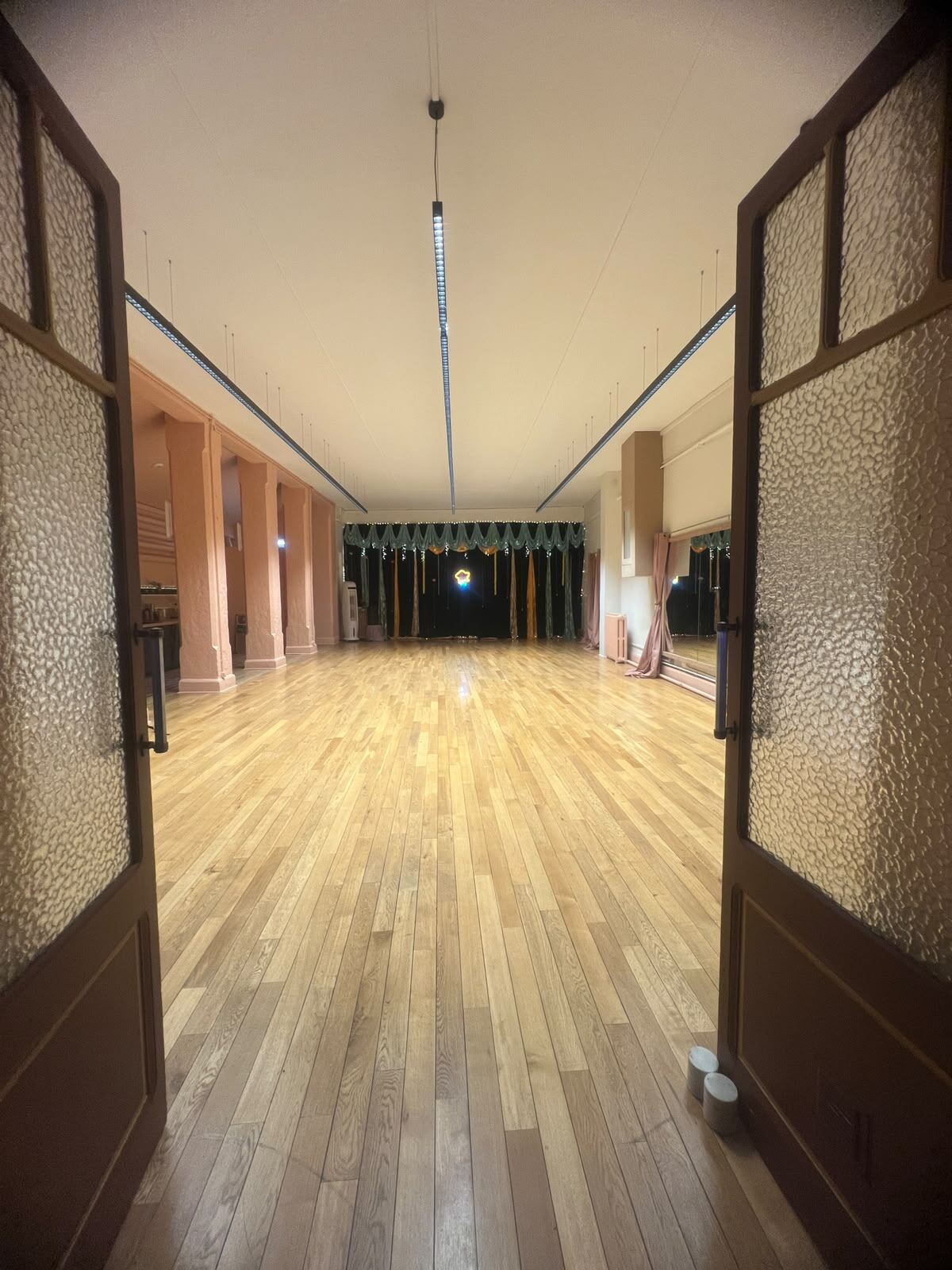 La salle vue depuis ses portes vitrées ouvertes : parquet en chêne clair, colonnes rosées et scène tendue de vert au fond